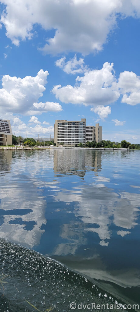 Disney's Contemporary Resort reflects in the calm water of a lake. The sky is blue with large, fluffy white clouds.