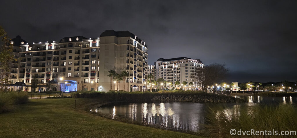 A nighttime view of Disney's Riviera Resort. The resort buildings are illuminated with warm lights, reflecting in the calm water of a lake.