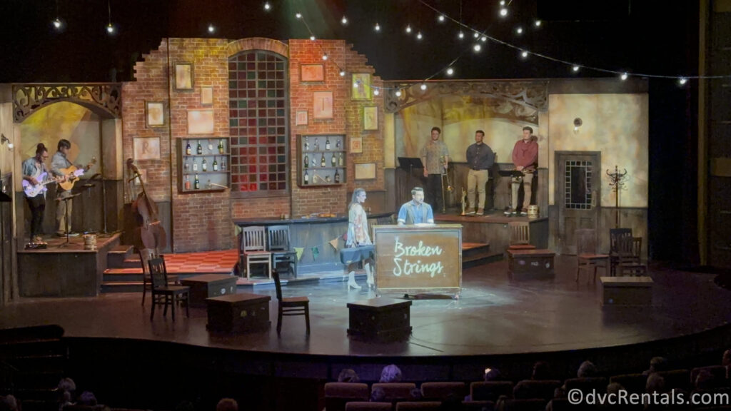 Two actors standing next to a piano that reads "Broken Strings" on the front of it.