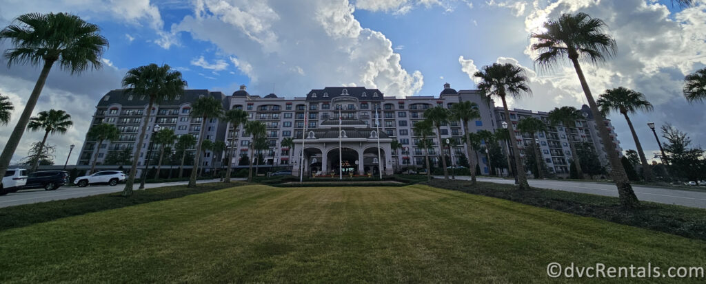 A wide-angle shot of Disney's Riviera Resort, showcasing its grand facade with a central archway and multiple balconies. Palm trees line the sides of the resort, and a blue sky with white clouds is visible above.