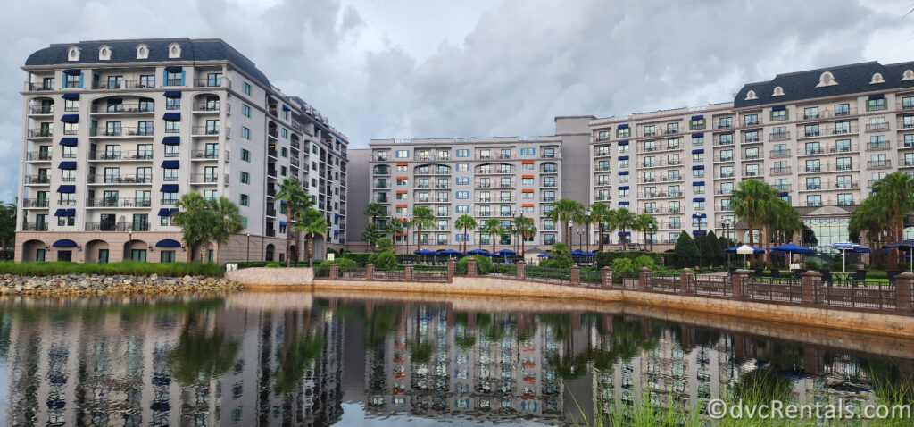 Disney's Riviera Resort. The resort is reflected in a large lake, showcasing its Mediterranean-inspired architecture with pastel colors and balconies.