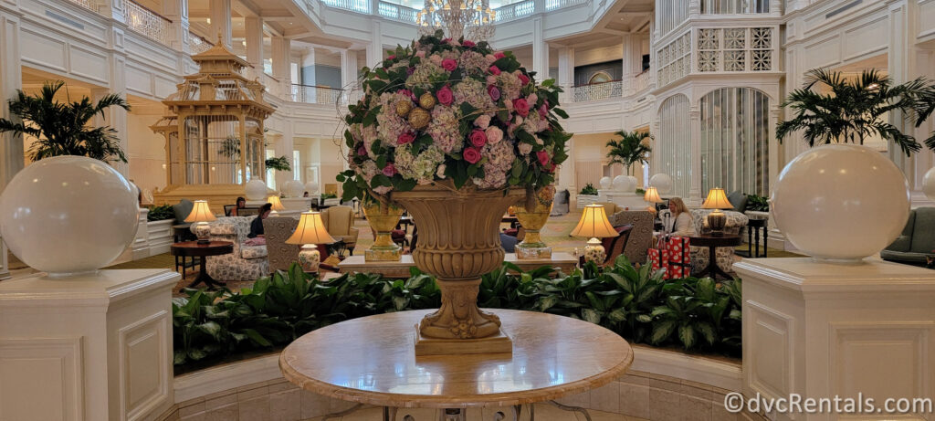 Grand Floridian Resort & Spa lobby. A large, ornate vase filled with colorful flowers sits in the center of the room, surrounded by potted plants and marble columns.