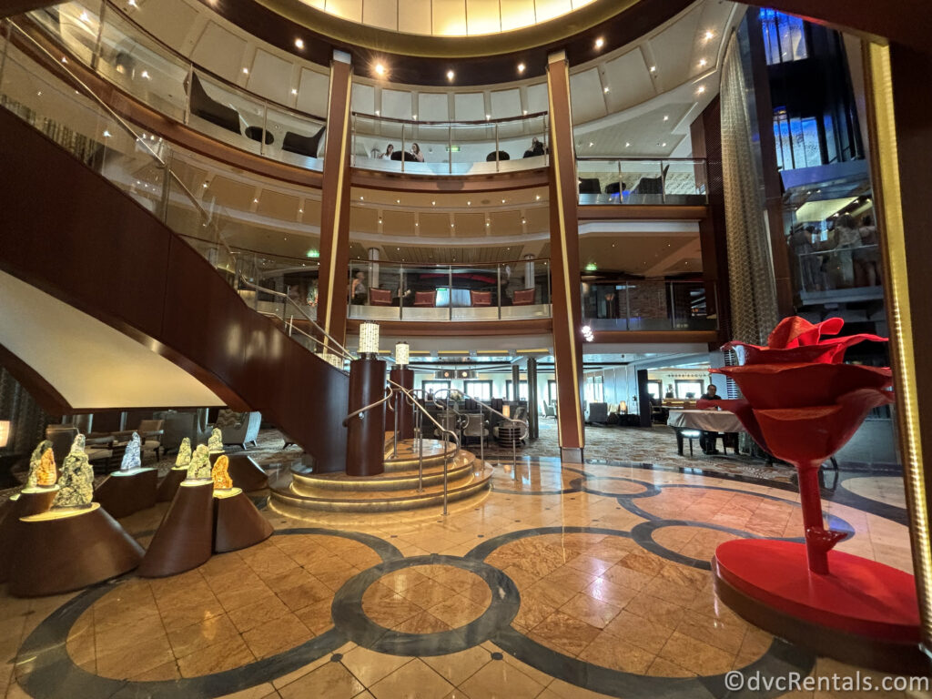 The interior of the Celebrity Reflection features a grand, multi-story atrium with staircases, glass railings, and polished marble floors. The space is decorated with art pieces, including a large red rose sculpture.