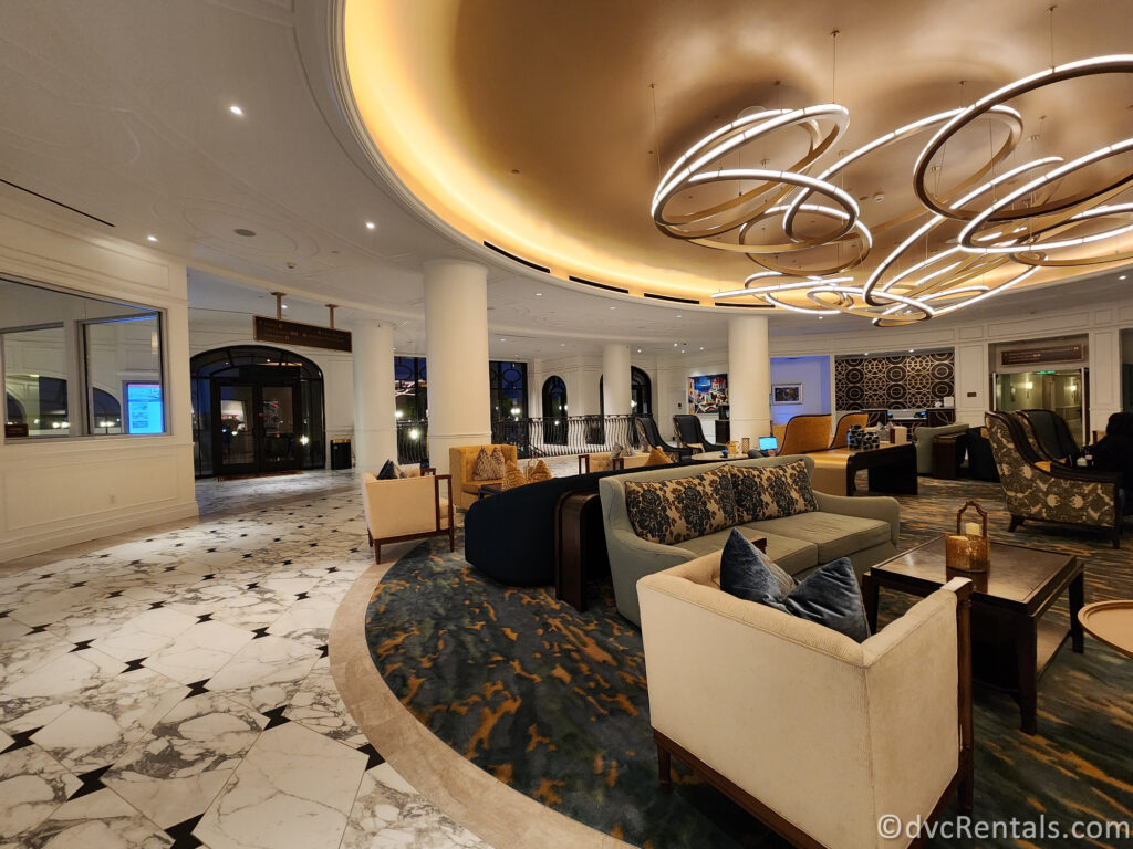 The lobby of Disney's Riviera Resort. It features a circular seating area with comfortable armchairs and sofas, a patterned rug, and a large, unique light fixture hanging from the ceiling.