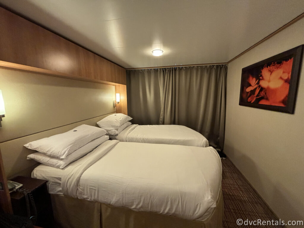 A cruise ship cabin with two single beds arranged side by side, each with white linens and multiple pillows. The room has warm wooden accents, soft lighting from wall-mounted lamps, and a piece of art featuring red flowers hanging on the wall.