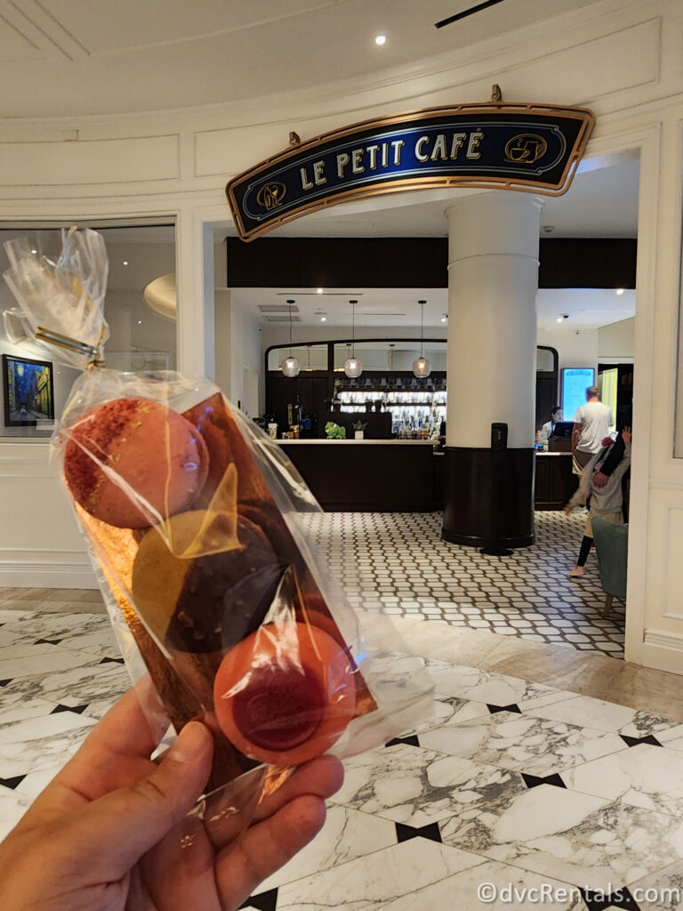 A person holds a small, clear bag of colorful macarons in their hand. In the background, the entrance to Le Petit Cafe at Disney's Riviera Resort is visible, with a bar and seating area in view.