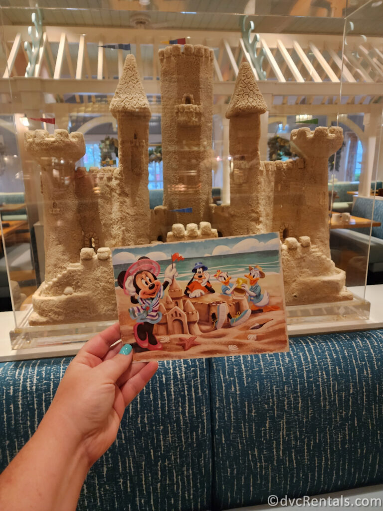 A woman holds a signature postcard from Cape May Cafe depicting Mickey Mouse, Minnie Mouse, Donald Duck, and Goofy building a sandcastle on a beach.