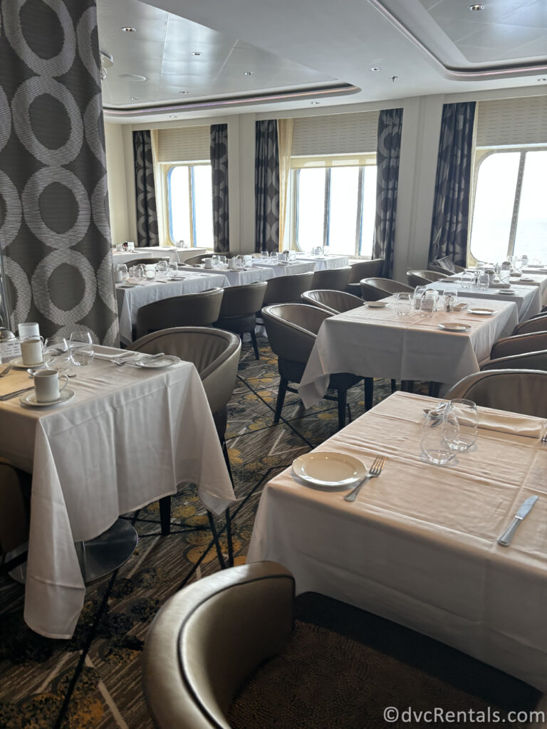 Interior of the Main Dining Room onboard the Celebrity Reflection. Tables covered in white linens in a chrome and silver dining room with gray chairs around the tables.