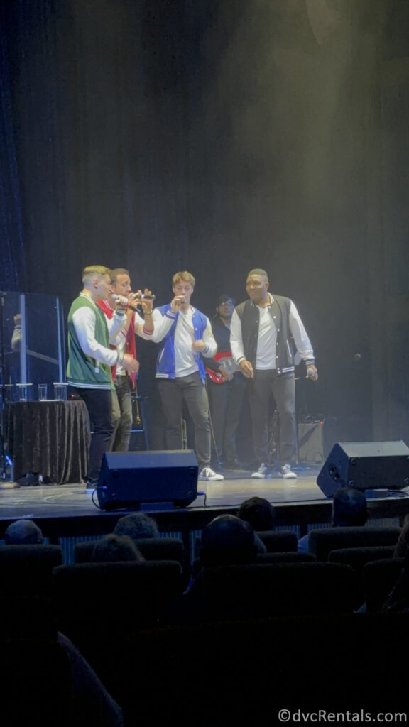 Four men standing on a stage in different colored shirts singing into microphones.