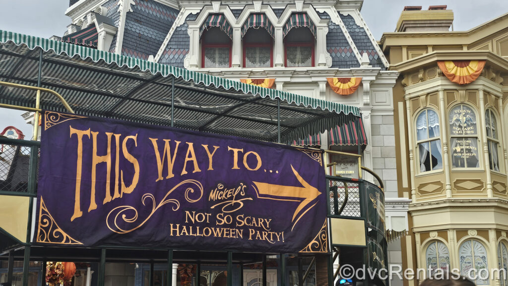 Purple sign on green double-decker bus with orange text reading “this way to Mickey’s Not So Scary Halloween Party!” with a right-facing arrow. Grey and brown Main Street U.S.A. buildings with orange, yellow, and brown Halloween decorations are in the background.