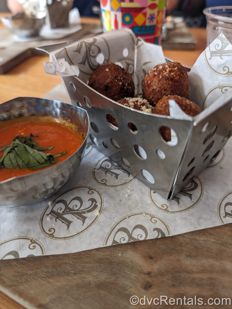 Seasonal Arancini from Bar Riva at Disney’s Riviera Resort. The arancini are served in a metal basket with a small metal bowl of marinara sauce for dipping, with white parchment paper underneath decorated with the golden “RR” logo of the resort.