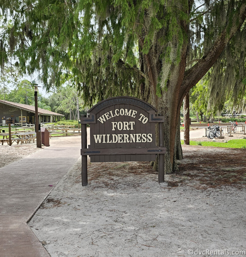 A wooden sign that reads "WELCOME TO FORT WILDERNESS" is located under a large tree with hanging moss. The sign is surrounded by greenery, including trees and shrubs, and there is a paved path leading up to it.