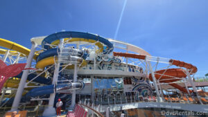 The blue, yellow, clear, and orange waterslide tubes on Royal Caribbean’s Utopia of the Seas under a sunny, blue sky. Orange and pink lounge chairs are also visible, as is a lifeguard and glass windows of the ship.