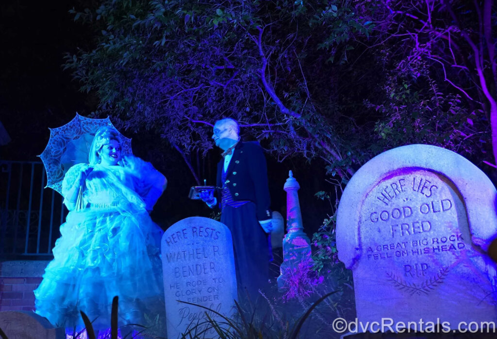 Cast Members in ghostly costumes amongst the tombstones in the queue for Haunted Mansion at Magic Kingdom. One is a female in a Victorian-style white, frilled ballgown with a parasol, and the other is male, dressed in a dark butler uniform.