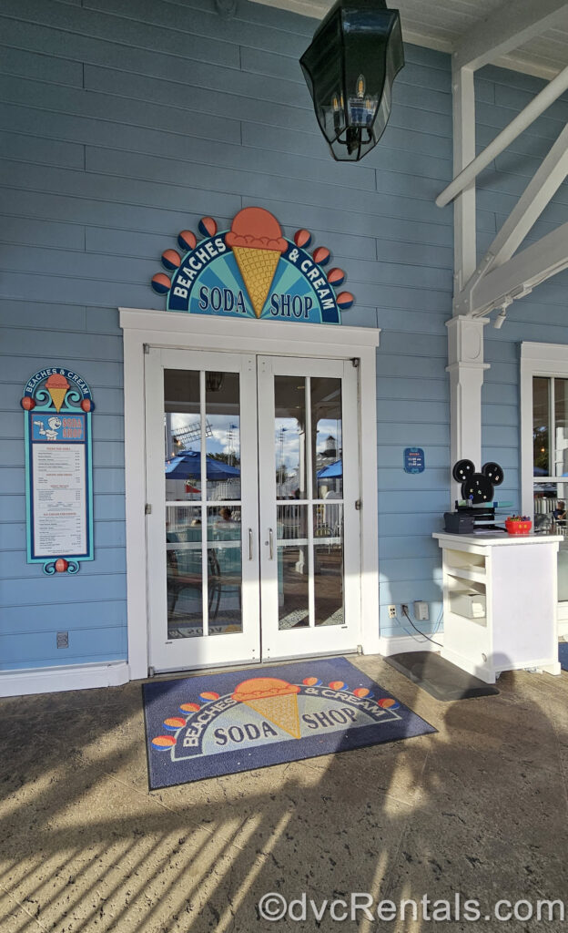 The white and glass double door entry way to Beaches & Cream Soda Shop at Disney’s Beach Club Villas. The building exterior is blue, and the restaurant sign is light and dark blue with a pink and brown ice cream cone. There is also a sign displaying the restaurant’s menu in the background.