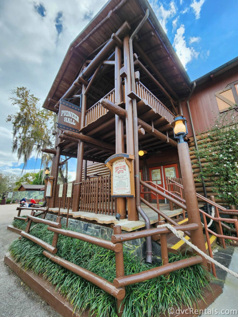 A wooden building with a large porch and a sign that says "PIONEER HALL". The building has a rustic appearance with exposed logs and a pitched roof. This is where Hoop-Dee-Doo takes place.