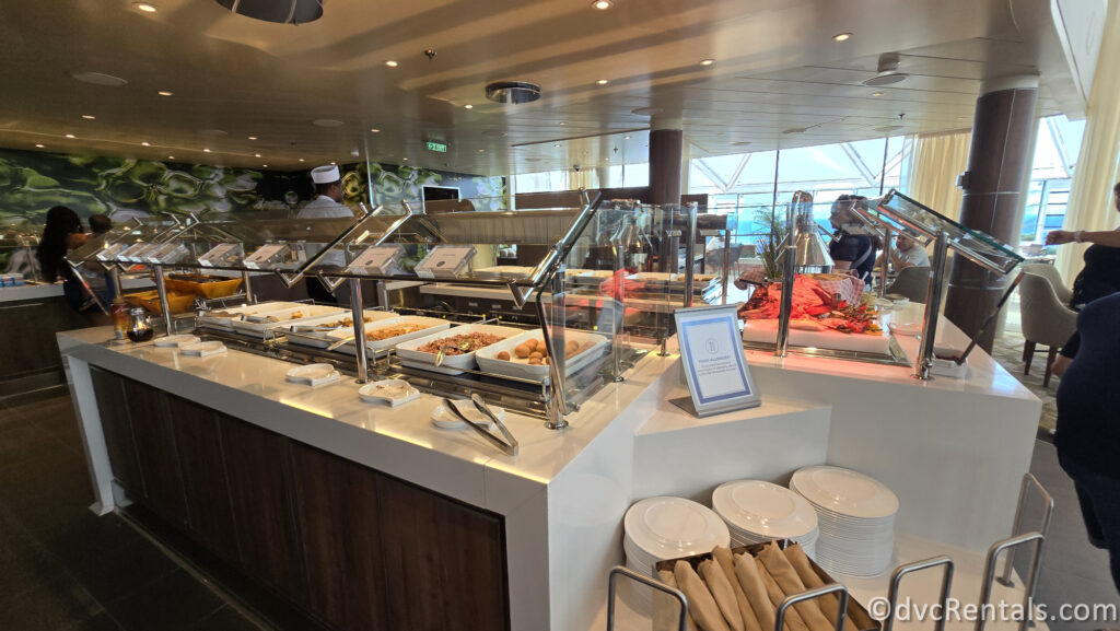 Various breakfast food offerings on the Windjammer buffet aboard Royal Caribbean’s Utopia of the Seas. There are white plates, white serving dishes, beige napkins, and glass coverings above the food. In the background, other guests and a chef are visible.