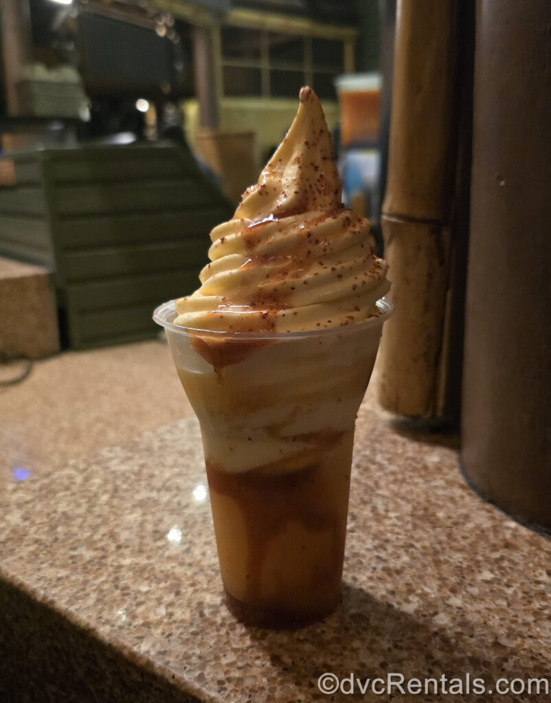 DOLE Whip Mango and Chamoy Float from Aloha Isle in Adventureland at Magic Kingdom. The treat is in a clear plastic cup with yellow juice at the bottom, topped with cream-colored swirled soft-serve DOLE Whip and spicy seasoning sprinkled over it.