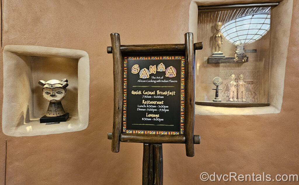 A dark wood sign reads “Sanaa” in colorful lettering. The sign displays the restaurant’s breakfast, lunch, dinner, and lounge hours, with a multicolored border and black background. Behind the sign there are multiple wooden African artifacts on display.