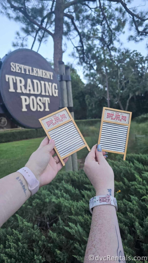 Two hands holding small washboard-shaped magnets in front of a wooden sign that reads "SETTLEMENT TRADING POST." The washboards have the words "HOOP DEE DOO REVUE" written on them.