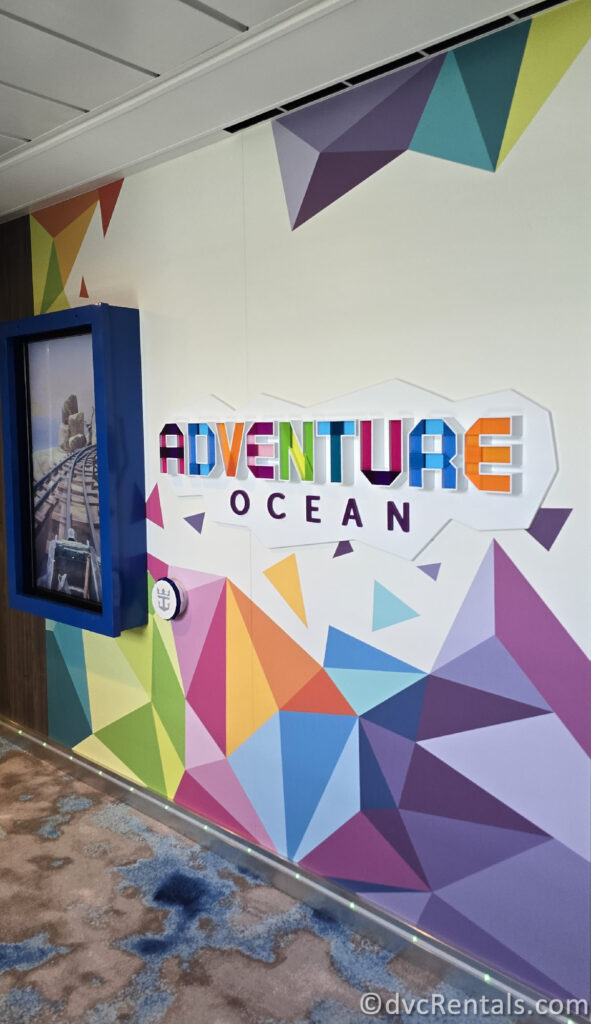 The interior, multi-color sign reading “Adventure Ocean” aboard Royal Caribbean’s Utopia of the Seas with a white background. On the wall above and below the sign there is a multi-color geometric mural with shades of purple, pink, blue, orange, yellow, and green.