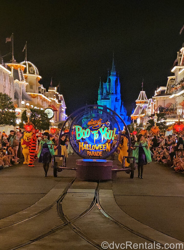 Looking toward Cinderella Castle down Main Street U.S.A. at night with a parade float displaying a sign reading “Mickey’s Boo-to-You! Halloween Parade”. There are Cast Members in green cloaks on either side of the float and crowds on each side of Main Street. The streetlights glow white, and Cinderella Castle is illuminated blue in the background.