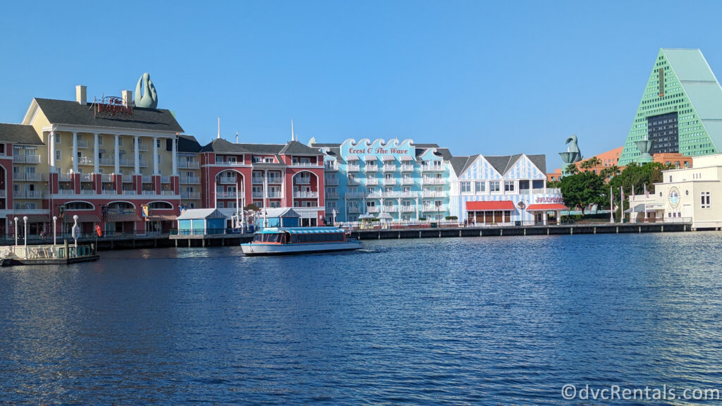 Disney’s BoardWalk Villas at Walt Disney World with Crescent Lake in the foreground. The building is colored in pastel shades of yellow, blue, white, and coral. There is a blue and white Friendship Boat on the water in front of the BoardWalk, and the green roof of the Swan & Dolphin resort can be seen in the background.