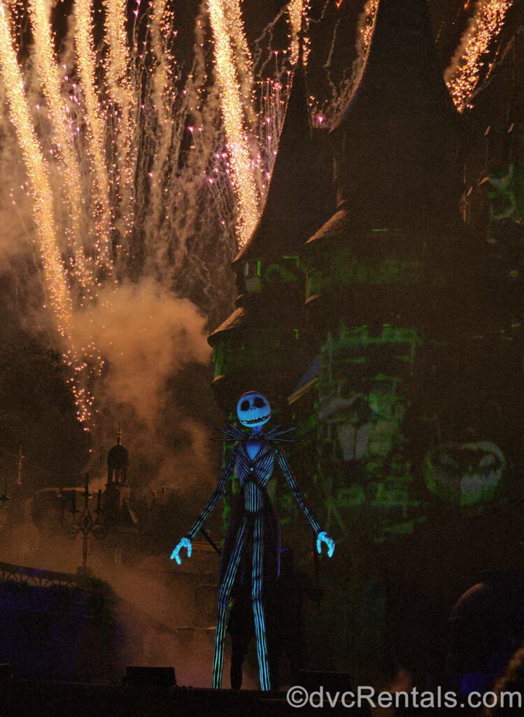 Golden fireworks erupt at the side of Cinderella Castle which is made to look green and spooky with special effects. A larger-than-life, black and white Jack Skellington puppet is performing in front of the fireworks.