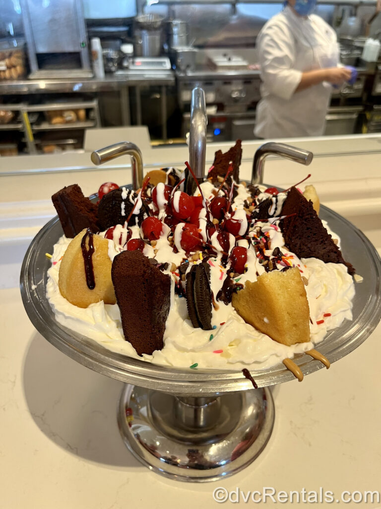 The large Kitchen Sink ice cream sundae is shown in a large, silver, sink-shaped bowl. There are vanilla and brownie cake pieces, lots of whipped cream, cherries, sprinkles, and chocolate sauce topping the dessert.