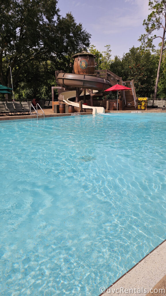 Swimming Pool at Fort Wilderness with clear blue water. A large, wooden water slide and a red umbrella can be seen in the background, along with lounge chairs and trees.