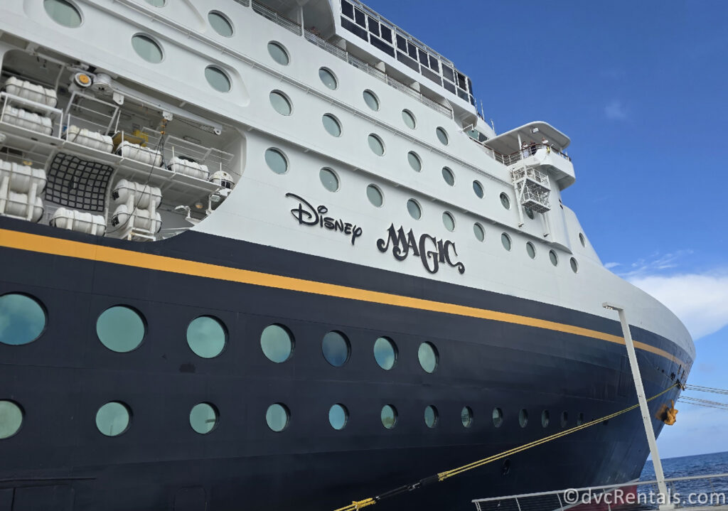 The white exterior of Disney’s Cruise Line’s Disney Magic ship under a blue sky. The bottom portion of the ship is navy-blue with a golden yellow stripe, and the words “Disney Magic” is written in large navy-blue letters along the side of the ship with water visible below.