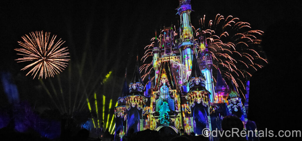 Yellow fireworks burst over Cinderella Castle at night, with neon green lasers point to the sky behind them. Cinderella Castle is covered in colorful Haunted Mansion themed projections including a ghost playing a pipe organ.