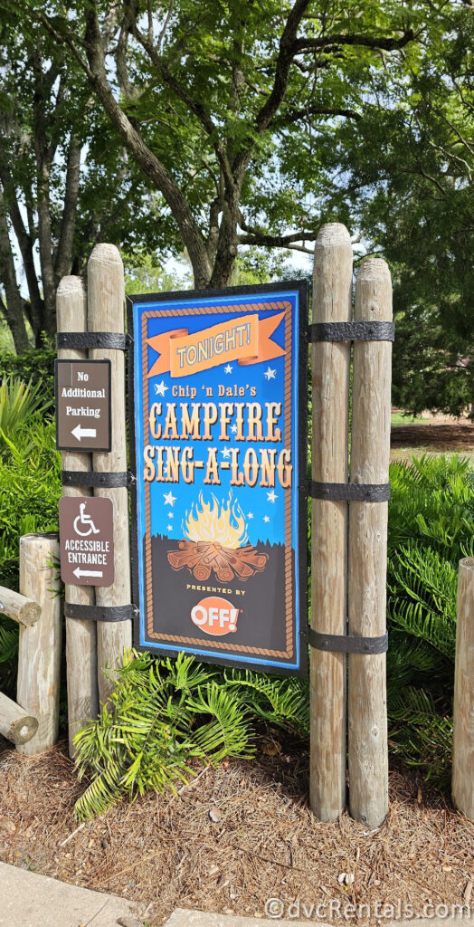 A wooden sign with a blue background and white lettering announcing "TONIGHT! Chip 'n Dale's CAMPFIRE SING-A-LONG PRESENTED BY OFF!" The sign is surrounded by trees and greenery.