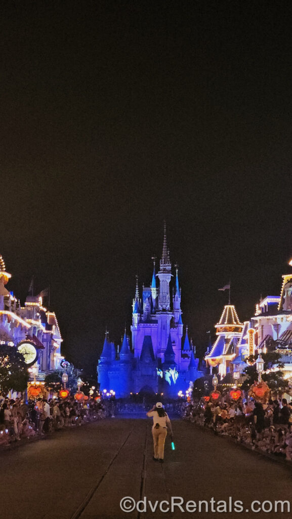 Looking down Main Street U.S.A. at night with streetlights aglow towards Cinderella Castle which is illuminated blue in the background. Main Street is clear down the middle with a Cast Member walking with a green glow stick, and crowds on either side ready for the parade to begin.