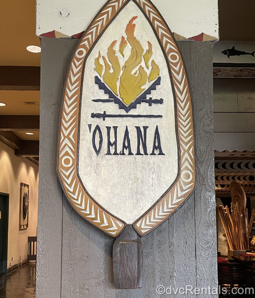 A paddle-shaped wooden sign reading “’Ohana” in dark blue writing. The sign is hung on a dark wood post and has yellow flames above the eatery’s name, with a light brown and white carved design around the edge.