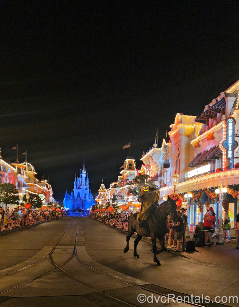 The Headless Horseman in a brown cloak holding a glowing jack-o-lantern trots down Main Street U.S.A. on a dark brown horse at night with streetlights aglow with Cinderella Castle, which is illuminated blue, in the background. Crowds view from either side of Main Street which is lit up with orange special lighting effects.