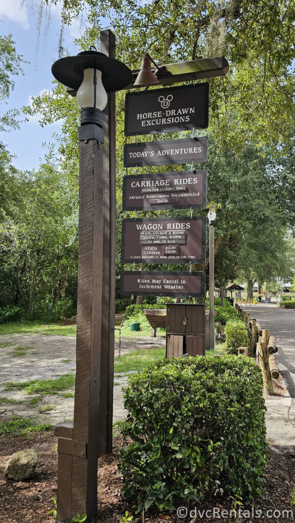 A wooden sign with a lamppost attached to it advertising horse-drawn excursions. The sign lists the available activities as carriage rides and wagon rides, along with prices for adults and children.