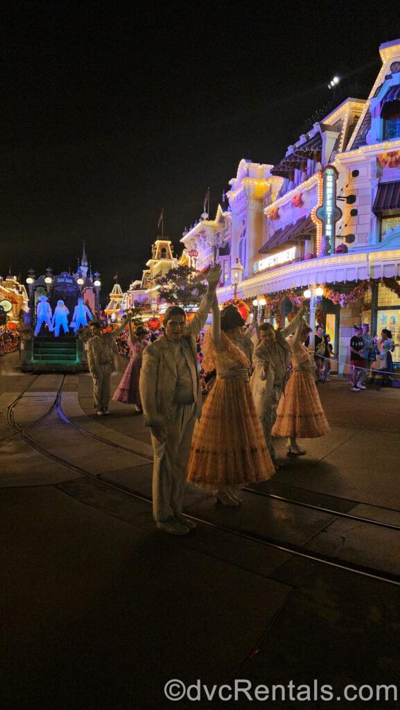 A group of parade performers dressed as ghostly ballroom dancers from Haunted Mansion pose on Main Street U.S.A. at night with a float carrying the 3 Hitchhiking Ghosts from Haunted Mansion behind them in the background.