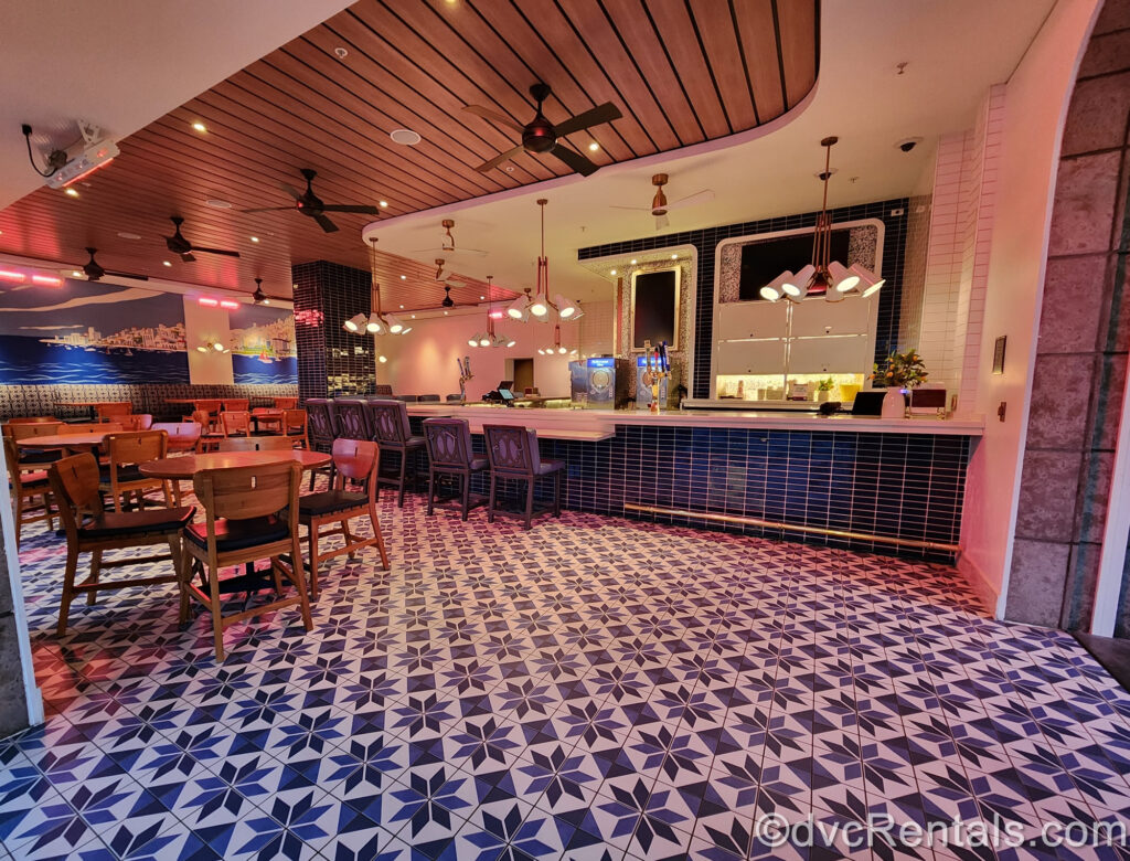 The interior of Bar Riva. The floor and bar are both decorated in blue and white patterns, with wooden tables. On the back wall there is a blue and white mural of the Mediterranean coastline and ocean, and blue seats line the bar.