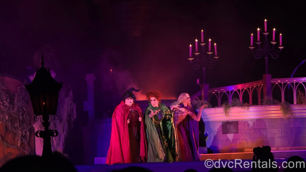 The Sanderson Sisters perform on stage. Mary has dark hair twirled upward and is wearing a red cloak, Winifred has red curly hair and a green cloak, and Sarah has long blonde hair and a purple cloak. There is purple and pink lighting, and 2 candelabras glow as fog swirls behind them.