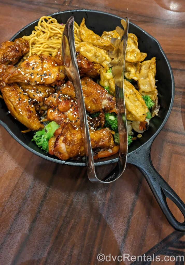 All-You-Care-To-Enjoy ‘Ohana Dinner Skillet. Honey-coriander chicken wings, dumplings, ‘Ohana noodles, and broccoli are served in a black skillet with silver tongs on top.