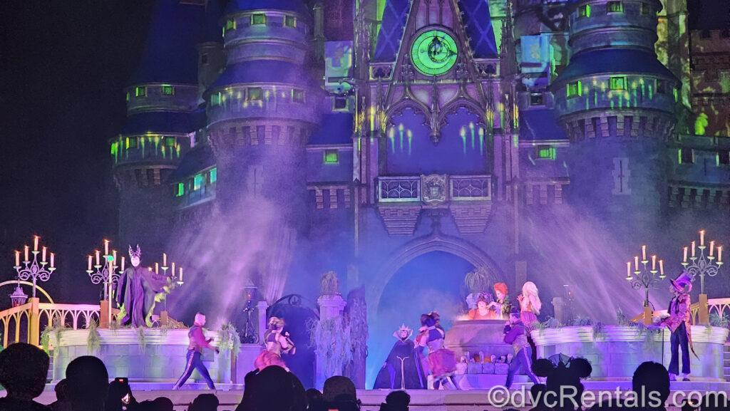 Maleficent, the Evil Queen, the Sanderson Sisters, and Dr. Facillier perform on stage with an ensemble of dancers in front of Cinderella Castle. The castle has spooky, neon green projections on it and a light fog rolls over the stage.