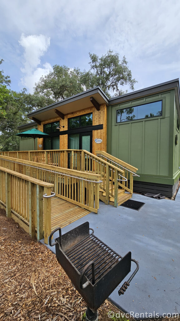 A green cabin with a wooden deck and ramp. There is a grill on the ground below the deck, and a patio umbrella can be seen on the deck.