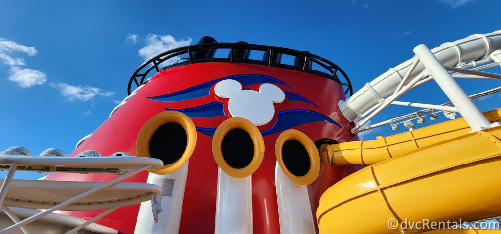 The bright red funnel of the Disney Wish. There is a white Mickey-head silhouette on the funnel with a royal blue wave design behind in. There are yellow and white tubular speakers for the ship’s horn visible in front of the funnel. To the right of the funnel there are white and yellow waterslide tubes visible, and a white canopy to the left.