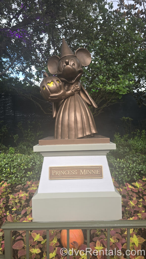 A copper-colored statue of Minnie Mouse in a princess costume holding a pumpkin with yellow Mickey-head silhouettes carved in it. There is green foliage in the background, and the sign at the base of the statue reads “Princess Minnie”.