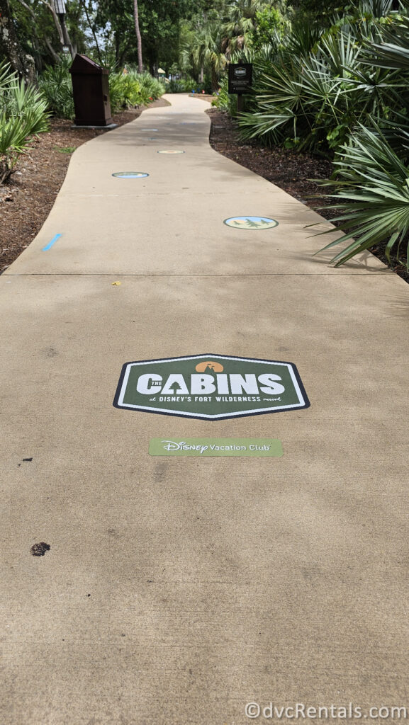 A paved walkway with decorative markers leading to a sign that reads "THE CABINS at Disney's Fort Wilderness Resort." The walkway is surrounded by trees and greenery.