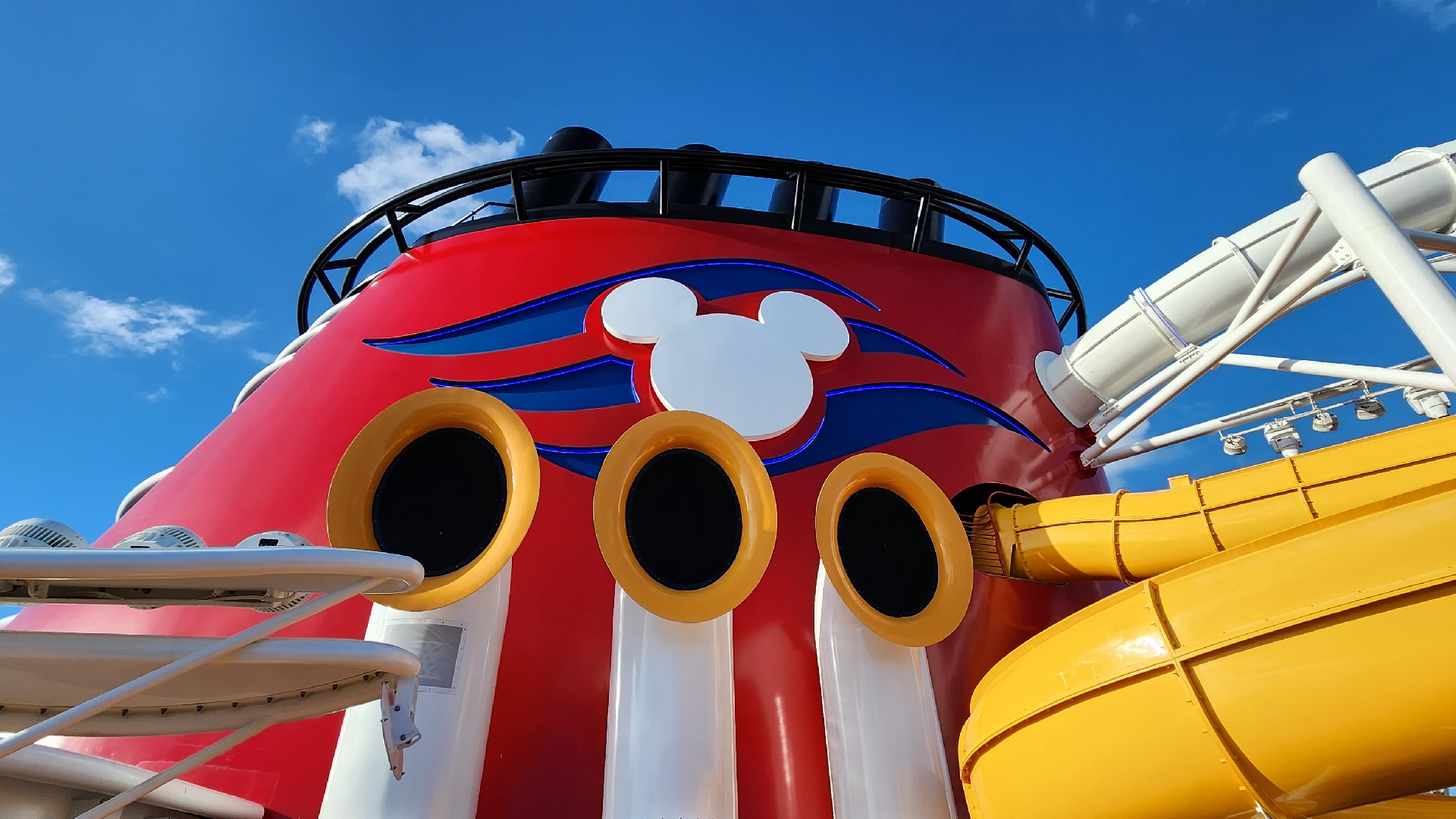 The bright red funnel of the Disney Wish. There is a white Mickey-head silhouette on the funnel with a royal blue wave design behind in. There are yellow and white tubular speakers for the ship’s horn visible in front of the funnel. To the right of the funnel there are white and yellow waterslide tubes visible, and a white canopy to the left.