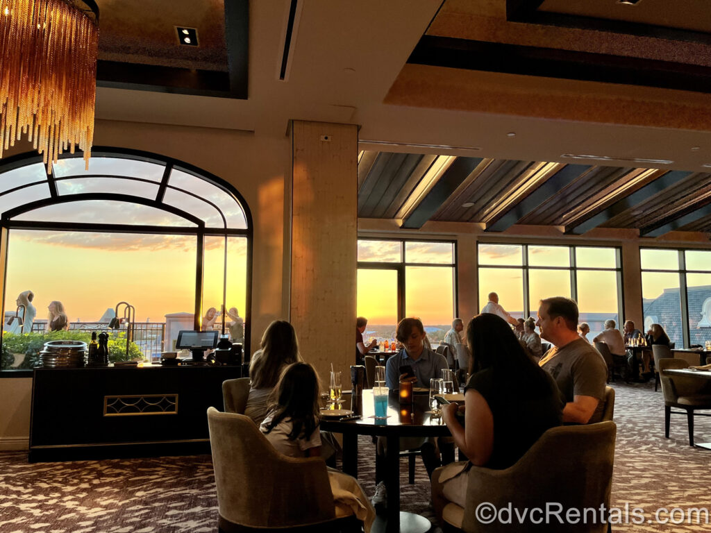 The light and dark brown dining room of Topolino’s Terrace at Disney’s Riviera Resort. Diners sit in around tables, and the orange and yellow light of sunset can be seen out of the restaurant’s large windows.