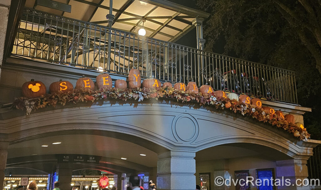 Several orange pumpkins carved to spell out “see ya real soon” with a Mickey-head at each end are lit up like jack-o-lanterns and displayed with seasonal leaf garland over the exit tunnel of Magic Kingdom Park.