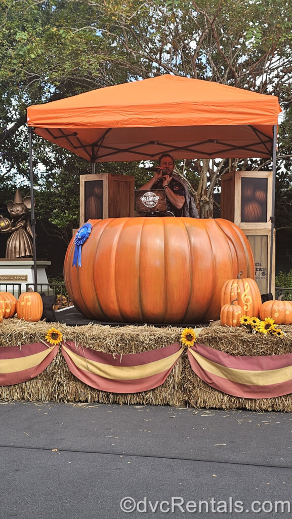 A DJ stands in a booth made to look like a larger-than-life orange pumpkin, under an orange tent. There is a blue prize ribbon on the large pumpkin, and it is set on a stage made of hay bales, with smaller pumpkins, red and yellow ribbons, and sunflowers around it.